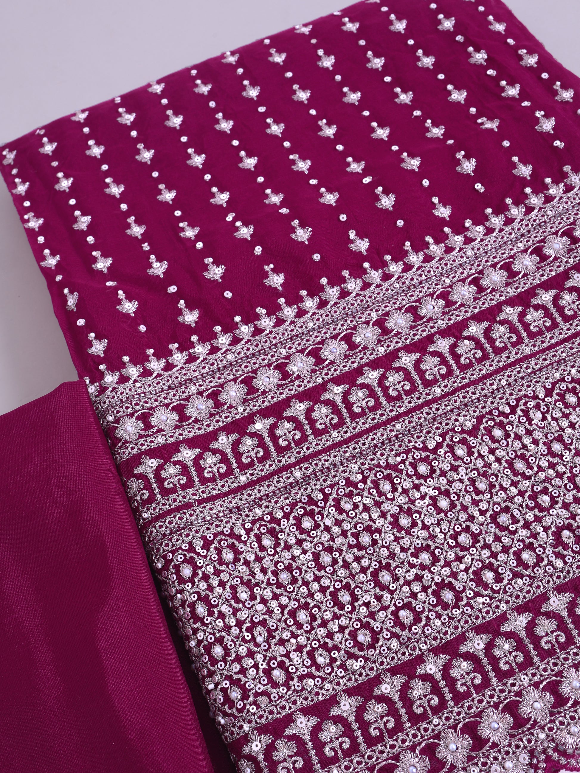 A close-up of a magenta pink fabric with intricate silver mirror work embroidery, showing detailed patterns and borders.