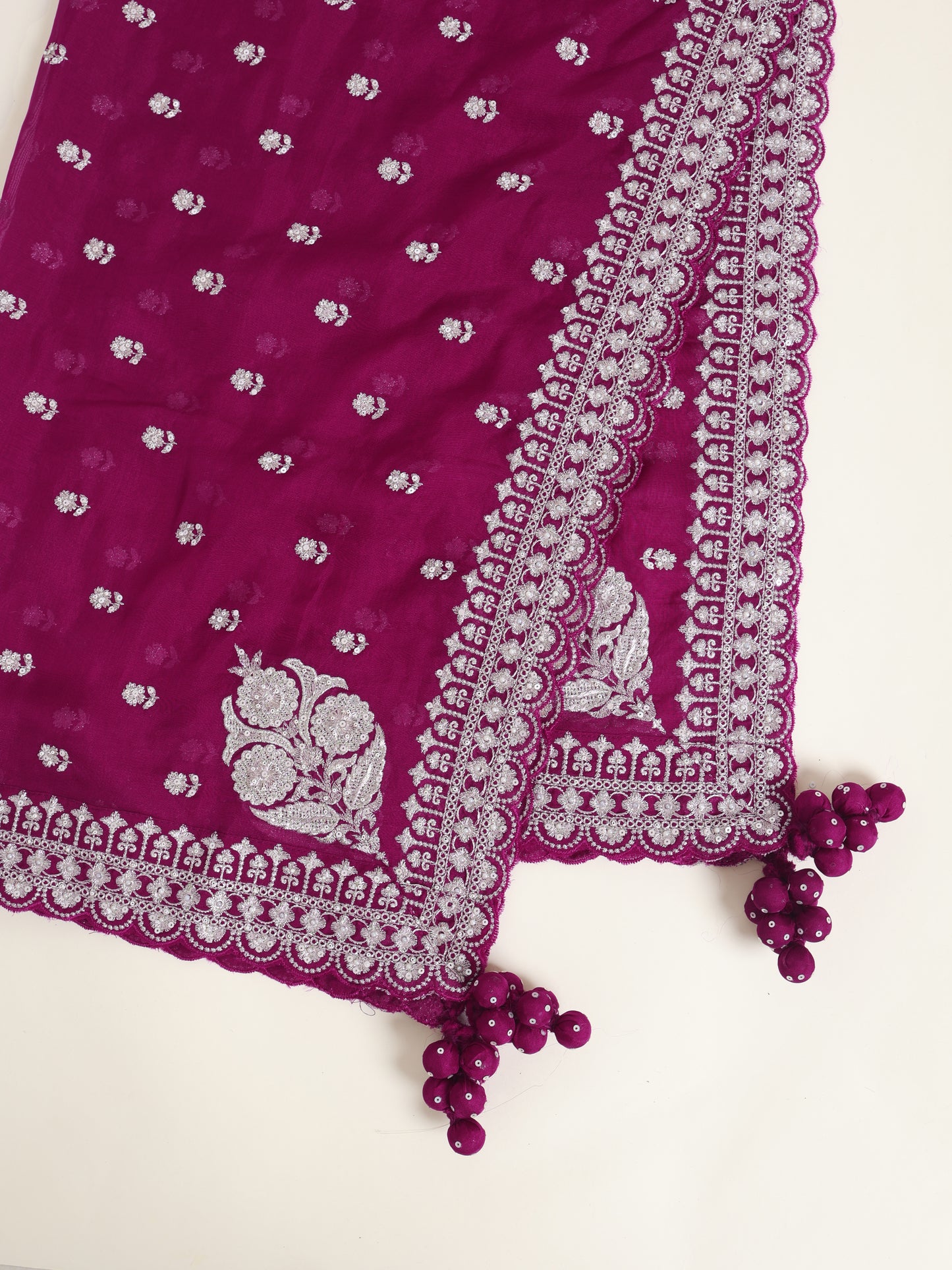 A deep magenta purple fabric, likely a dupatta or scarf, featuring small white floral prints, a large white floral motif near the corner, and an intricate white scalloped border. The fabric is styled diagonally on a light cream background with matching purple tassel beads.