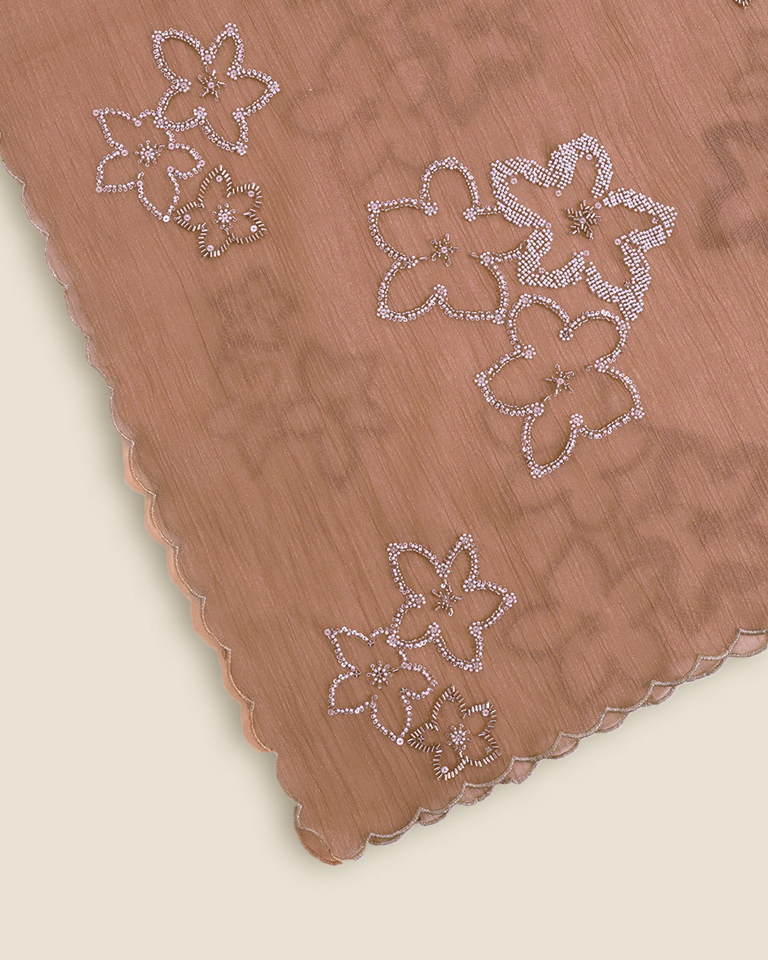 A close-up of a peach-colored fabric with scalloped edges and embroidered floral patterns embellished with small silver rhinestones, set against a light cream background.