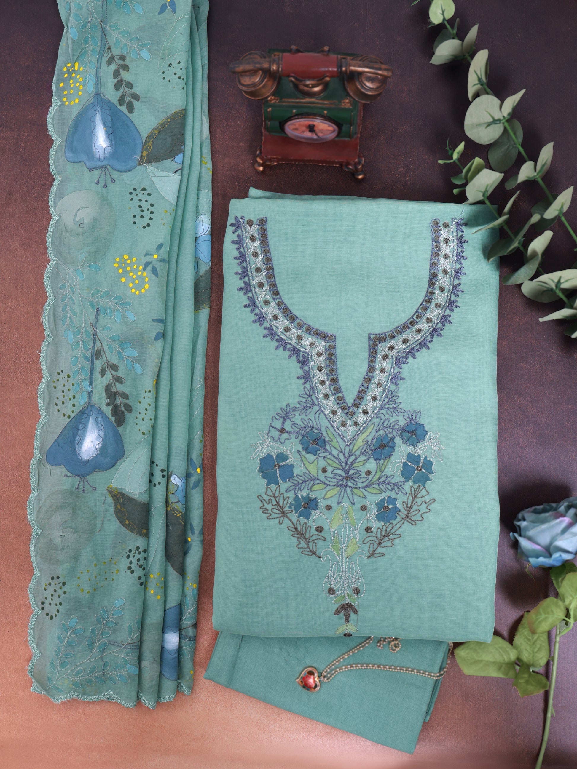 A folded mint green salwar suit set with floral embroidery, featuring a matching printed dupatta and decorative accessories, arranged on a brown surface.
