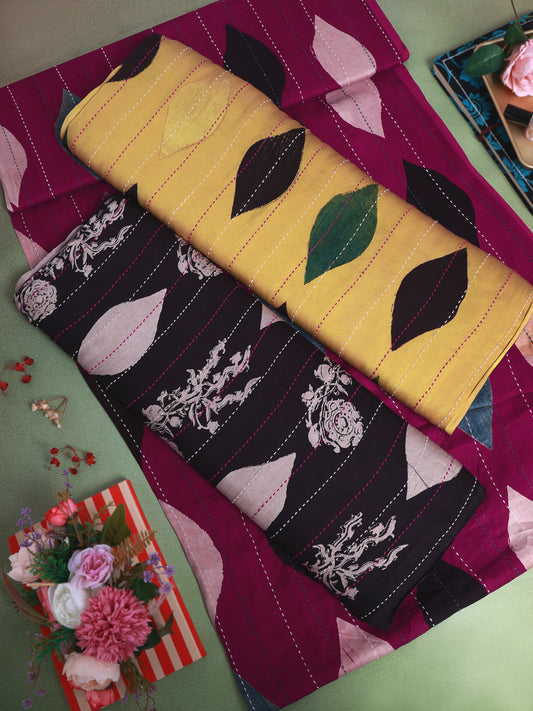 Three folded fabric pieces with distinct prints are arranged on a light green surface: a magenta fabric with pale pink leaf motifs, a mustard yellow fabric with dark green leaf motifs, and a black fabric with white floral and vine motifs. Decorative flowers and a striped gift box are placed around them.